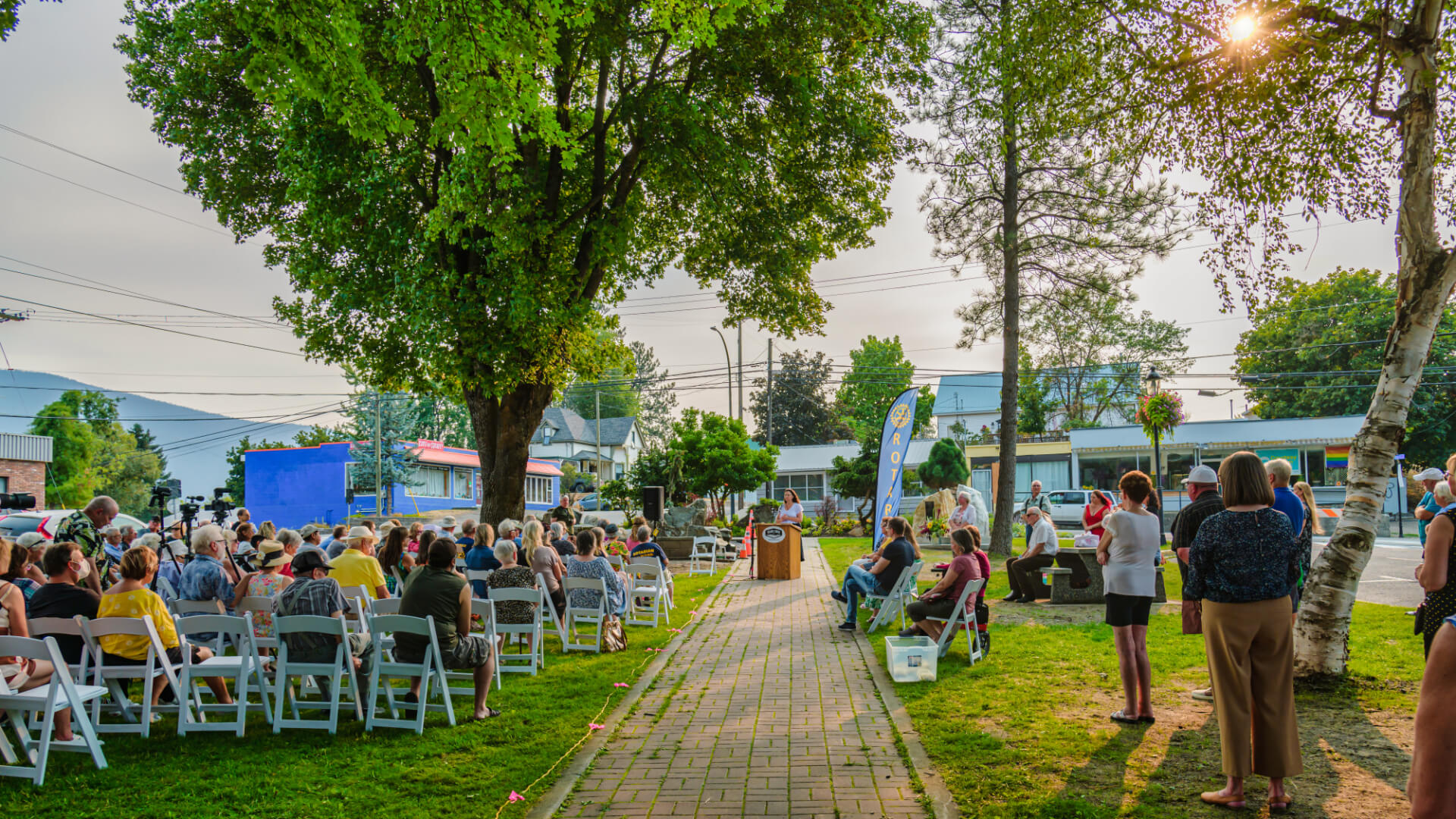 A crowd seated in rows of white folding chairs on a park lawn watching a speaker at a podium, with tents, parked cars and leafy trees along the street behind them