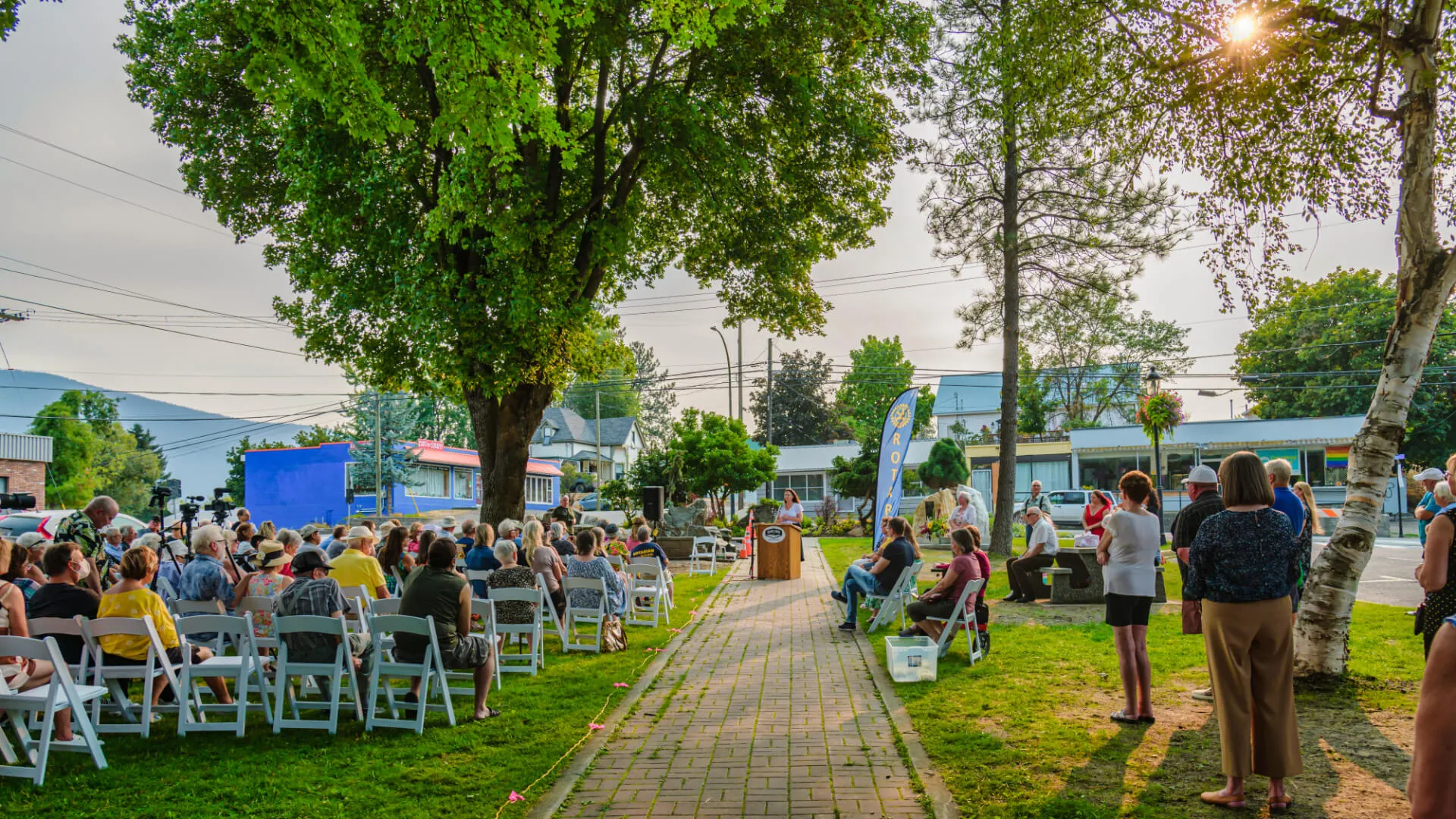 A crowd seated in rows of white folding chairs on a park lawn watching a speaker at a podium, with tents, parked cars and leafy trees along the street behind them