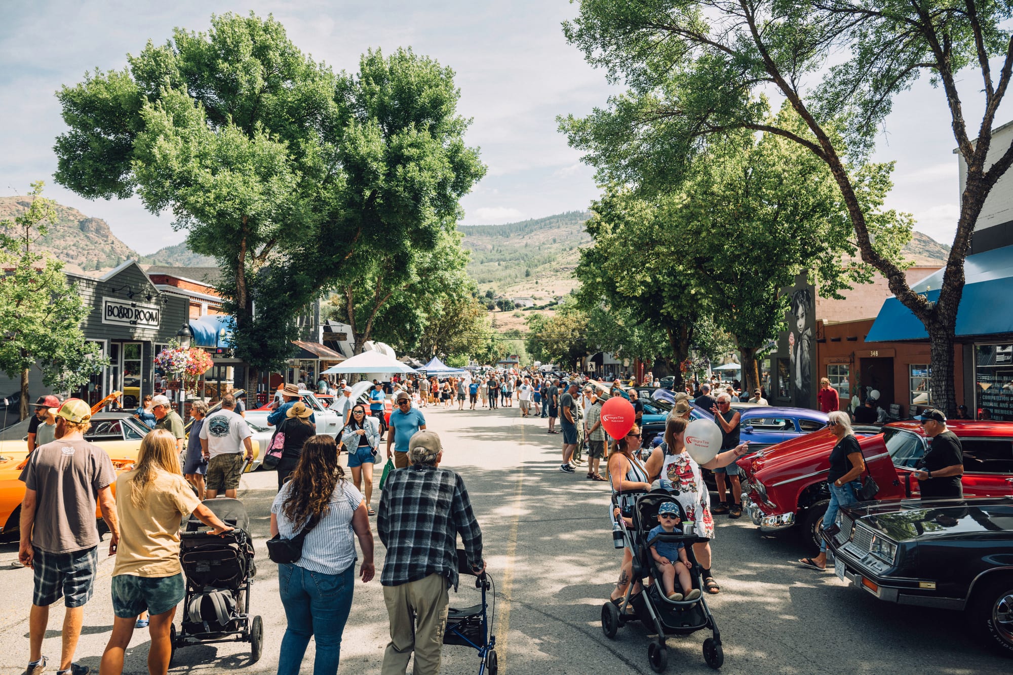 A downtown Grand Forks street packed with people and parked classic cars at a summer street festival, dry hills rising behind the storefronts