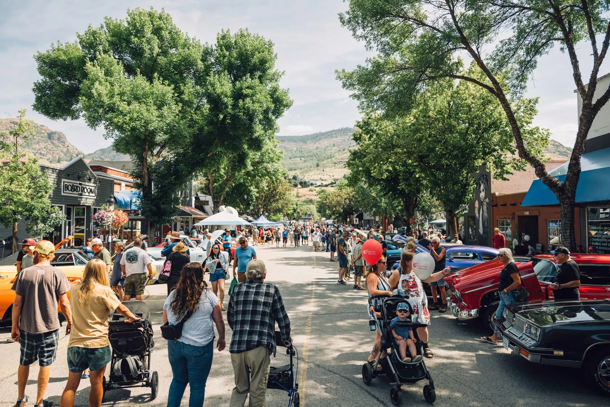 A downtown Grand Forks street packed with people and parked classic cars at a summer street festival, dry hills rising behind the storefronts
