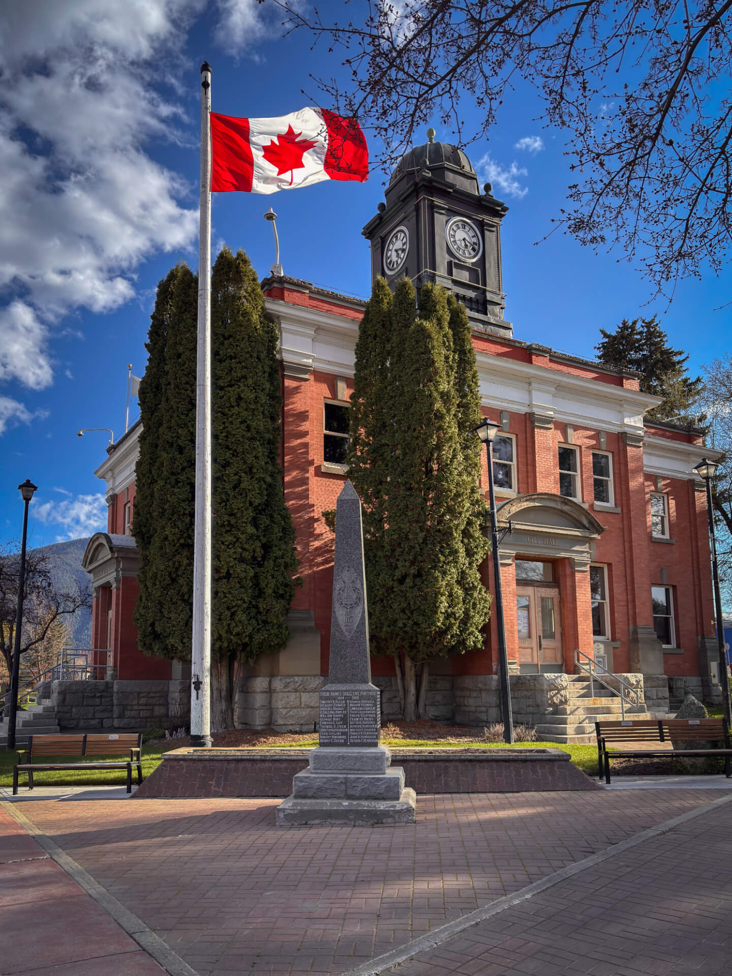 Grand Forks City Hall, a red-brick building with a black clock tower, behind a Canadian flag and the stone cenotaph on its brick plaza