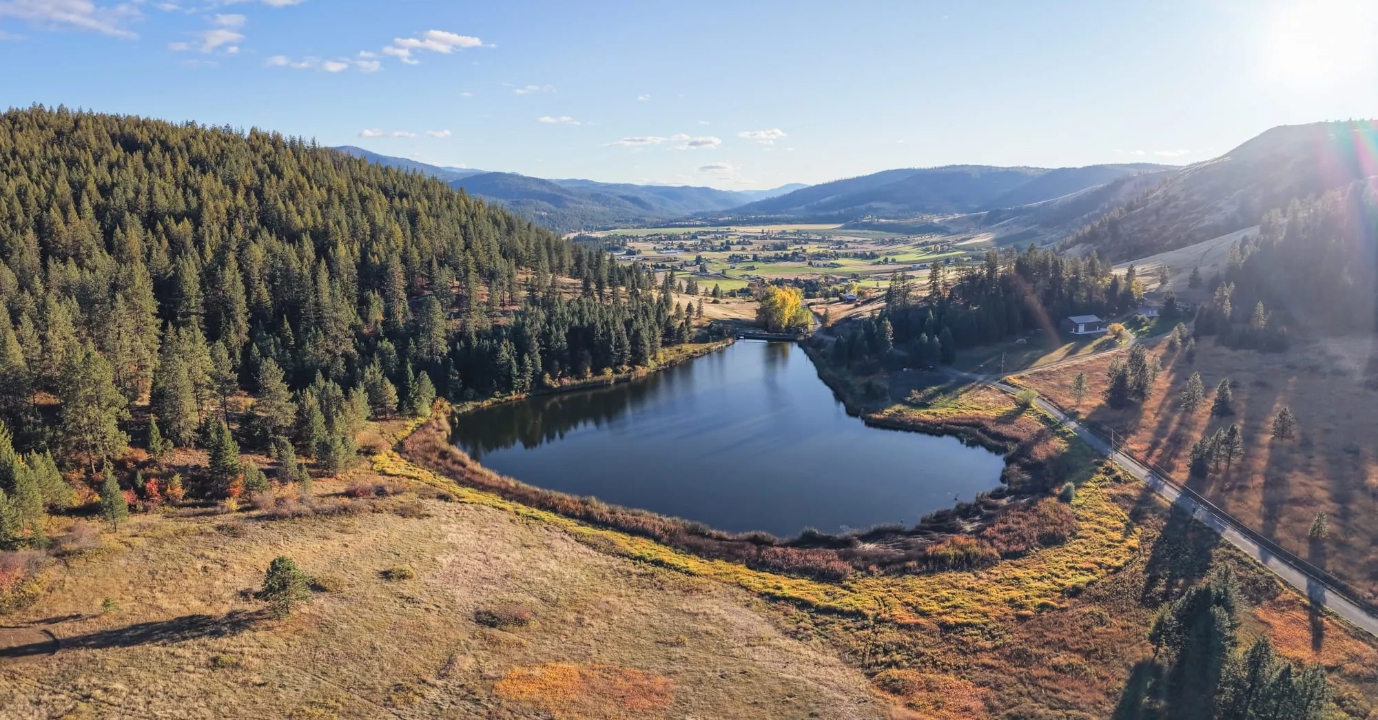 Aerial view of a small dark lake ringed by pine forest and autumn grassland, with farmland and layered blue mountains stretching down the valley beyond