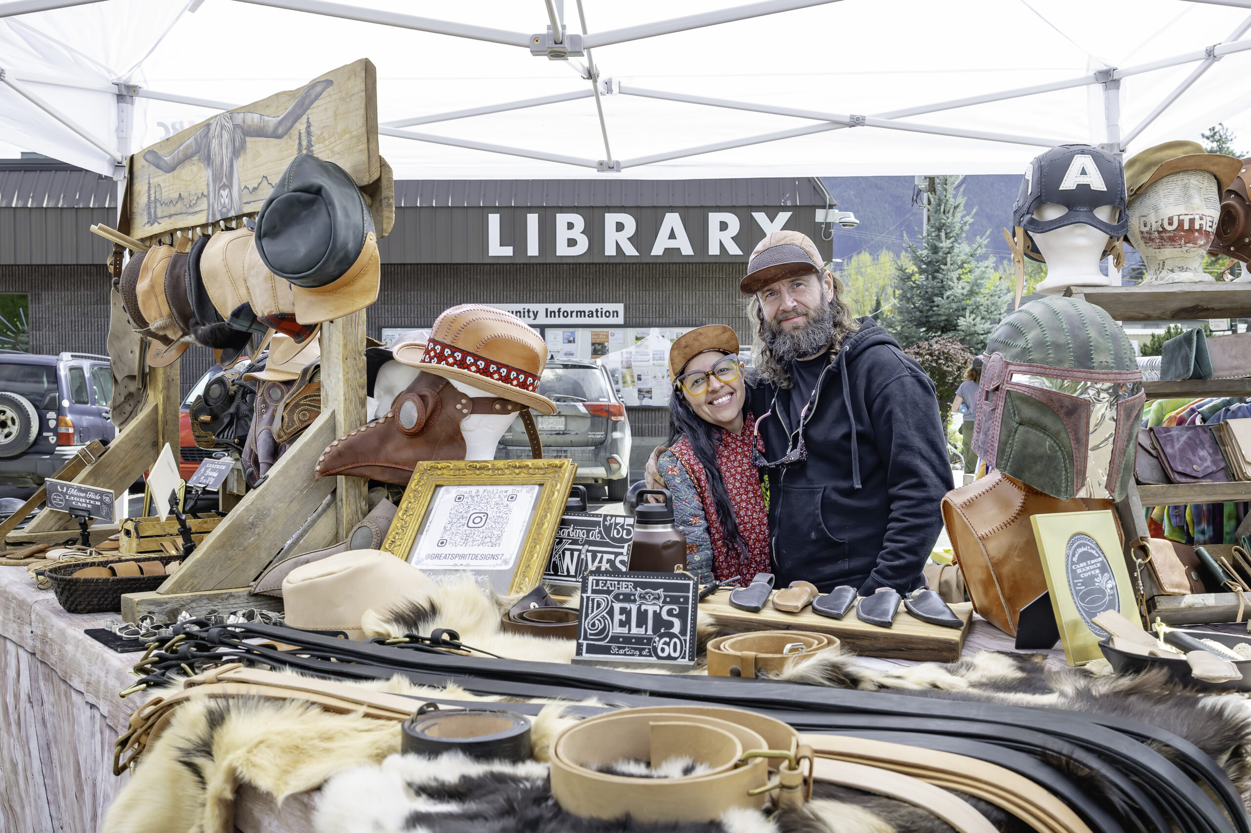 A leatherworking couple smiling behind their market stall of hats, belts and masks